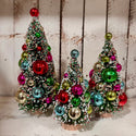 Three small frosted Christmas tree ornaments in different sizes, decorated with colorful glittery baubles in red, green, blue, pink, and gold, set against a rustic white wood plank background.