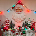 A large Santa Claus head wall hanging is displayed on a decorated Christmas mantel with smaller Santa figures, miniature trees, red and green ornaments, and multicolored string lights against a white paneled wall.