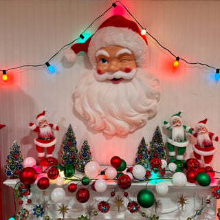 A large Santa Claus head wall hanging is displayed on a decorated Christmas mantel with smaller Santa figures, miniature trees, red and green ornaments, and multicolored string lights against a white paneled wall.