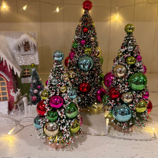 Three small artificial Christmas trees with white faux snow and colorful metallic ornaments, displayed on a tabletop with warm string lights and a decorative house in the background.