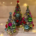 Three small artificial Christmas trees decorated with colorful shiny ornaments and white faux snow, arranged on a tabletop with warm string lights in the background.
