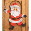 A vintage-style hanging Christmas door decoration featuring a smiling Santa Claus in a red suit with white fur trim, black boots and gloves, holding a candy cane, set against a wooden door background.