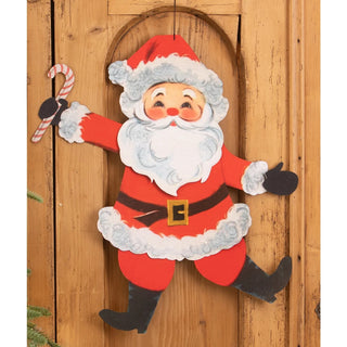 A vintage-style hanging Christmas door decoration featuring a smiling Santa Claus in a red suit with white fur trim, black boots and gloves, holding a candy cane, set against a wooden door background.
