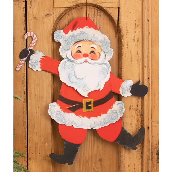 A vintage-style hanging Christmas door decoration featuring a smiling Santa Claus in a red suit with white fur trim, black boots and gloves, holding a candy cane, set against a wooden door background.