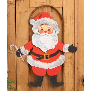A festive Santa Claus wooden door hanging decoration with a red suit, white beard and trim, black gloves and boots, and a candy cane, shown against a rustic wooden door background.