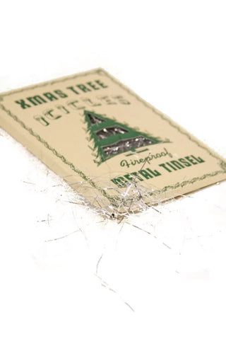 A square beige paper label or tag with green text reading 'XMAS TREE DECOR' and a green Christmas tree illustration, placed on a white background with scattered silver tinsel around it.
