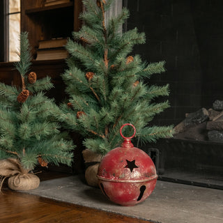A distressed red metal holiday bell with a star-shaped cutout sits on a fireplace hearth next to two small artificial Christmas trees in a rustic indoor setting.